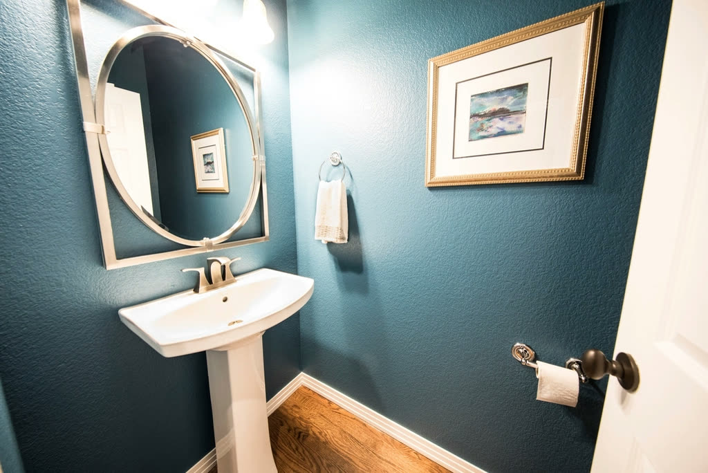 A small bathroom with deep teal walls, a white pedestal sink and a round mirror